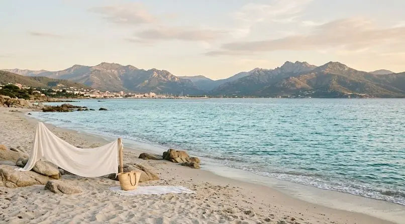 Rivage de Porticcio avec sable clair, mer calme et montagnes au loin, un drapé blanc et un sac en paille posés près des rochers