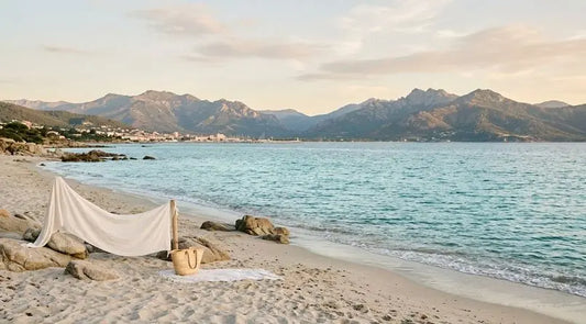 Rivage de Porticcio avec sable clair, mer calme et montagnes au loin, un drapé blanc et un sac en paille posés près des rochers